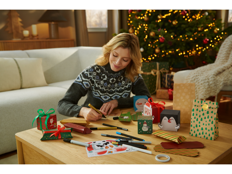 Frau bereitet Weihnachtsgeschenkverpackungen vor, mit Weihnachtsbaum im Hintergrund.