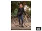 Frau in schwarzem Mantel und Jeans, hält ein Fahrrad in einem Park.