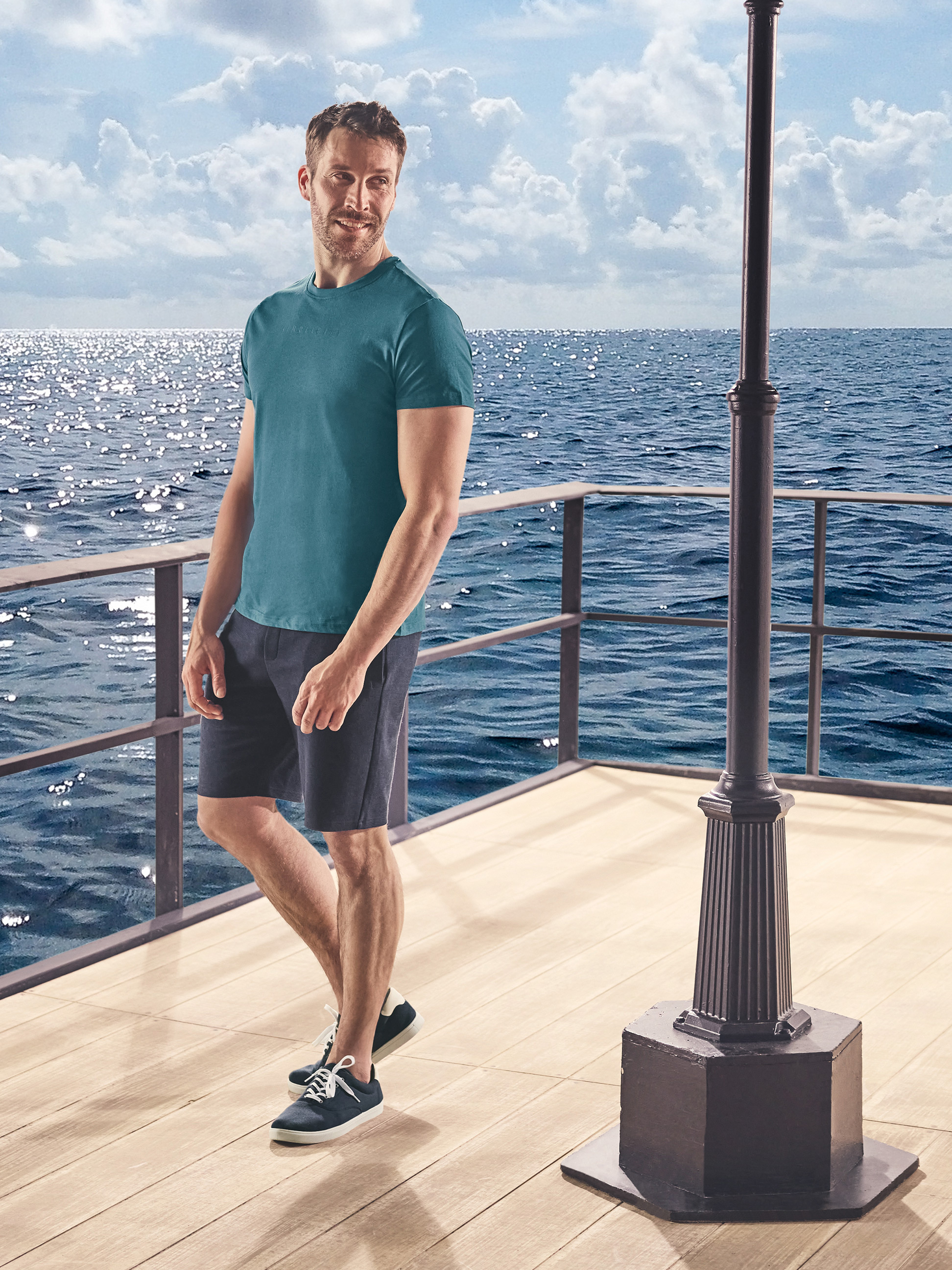 Mann in lässigem T-Shirt und Shorts auf einem Holzsteg am Meer.
