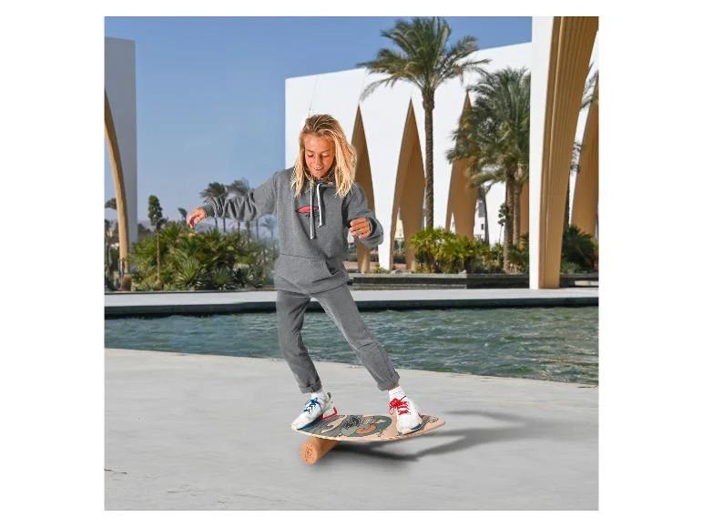 Blondes Kind in grauem Hoodie und Jogginghose balanciert auf einem Holz-Balanceboard.