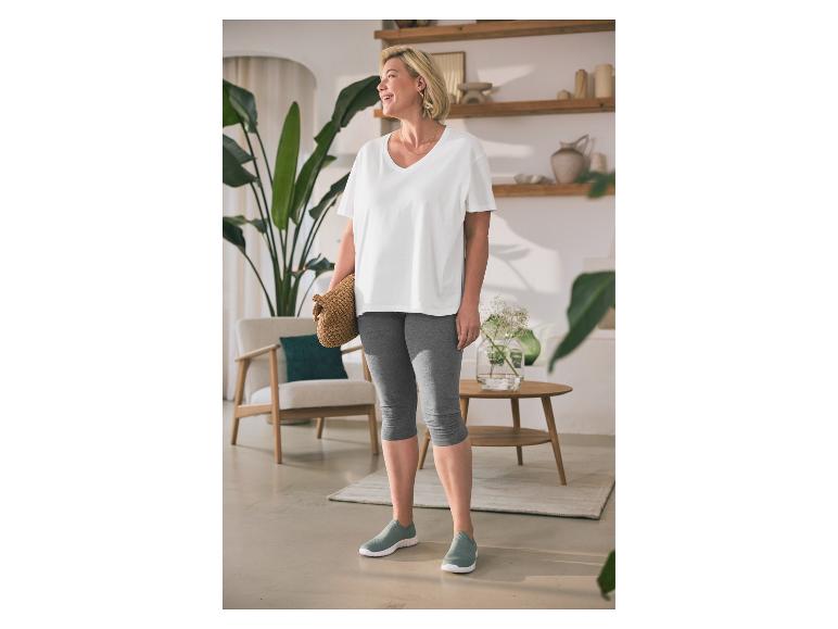 Frau in weißem T-Shirt und grauen Leggings mit Handtasche steht in gemütlichem Raum.