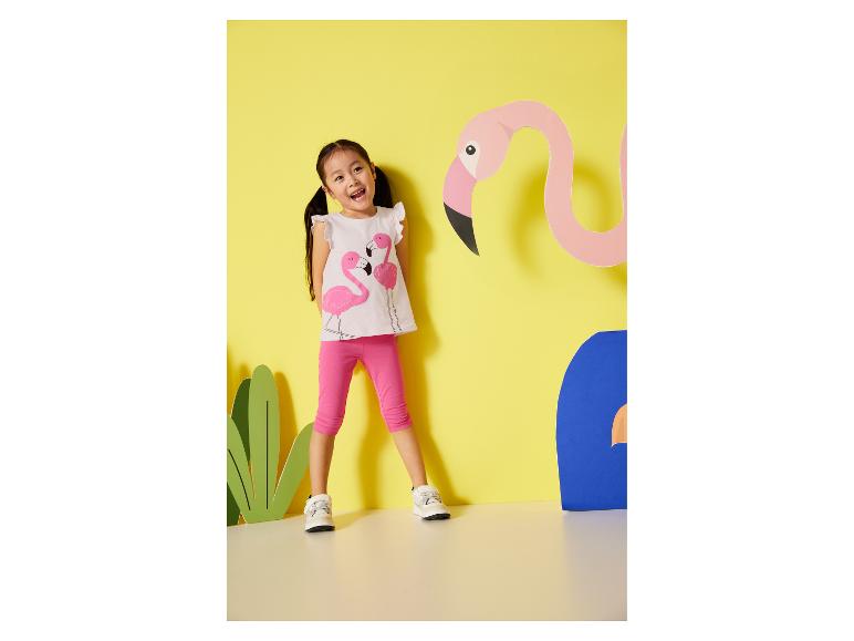 Ein Kind in rosa Leggings und einem Flamingo-Shirt posiert vor bunten Papierdekorationen.