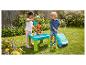 Zwei Kinder spielen mit einem Wasser- und Sandtisch, einer Seifenblasenmaschine und einem Aufsitzspielzeug im Garten.
