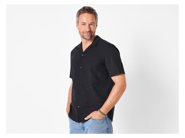 Mann in schwarzem Kurzarmhemd und Jeans, posiert mit Händen in den Taschen.