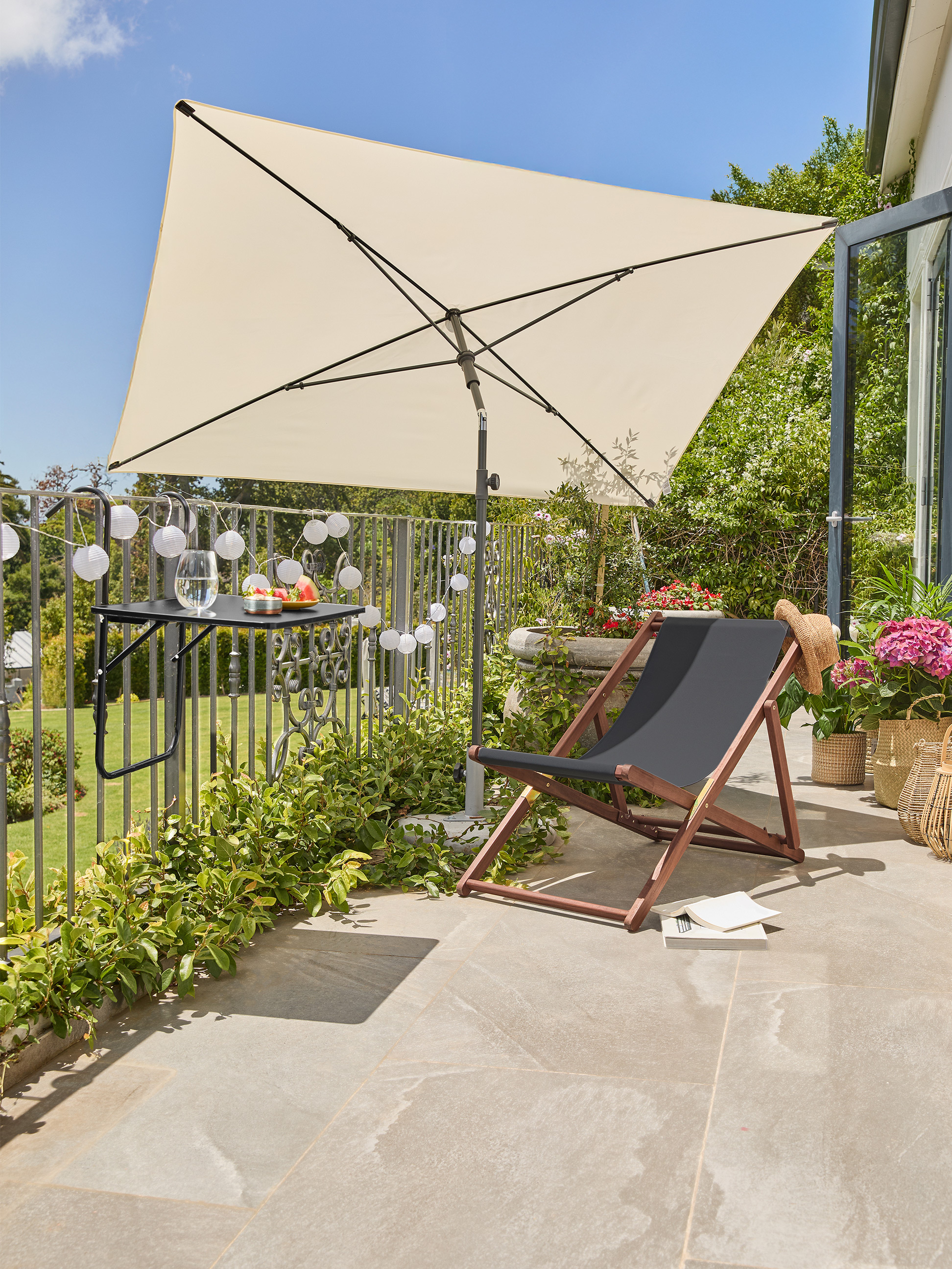 Balkon mit beigem Sonnenschirm, schwarzem Liegestuhl und Tisch mit Getränken und Obst.