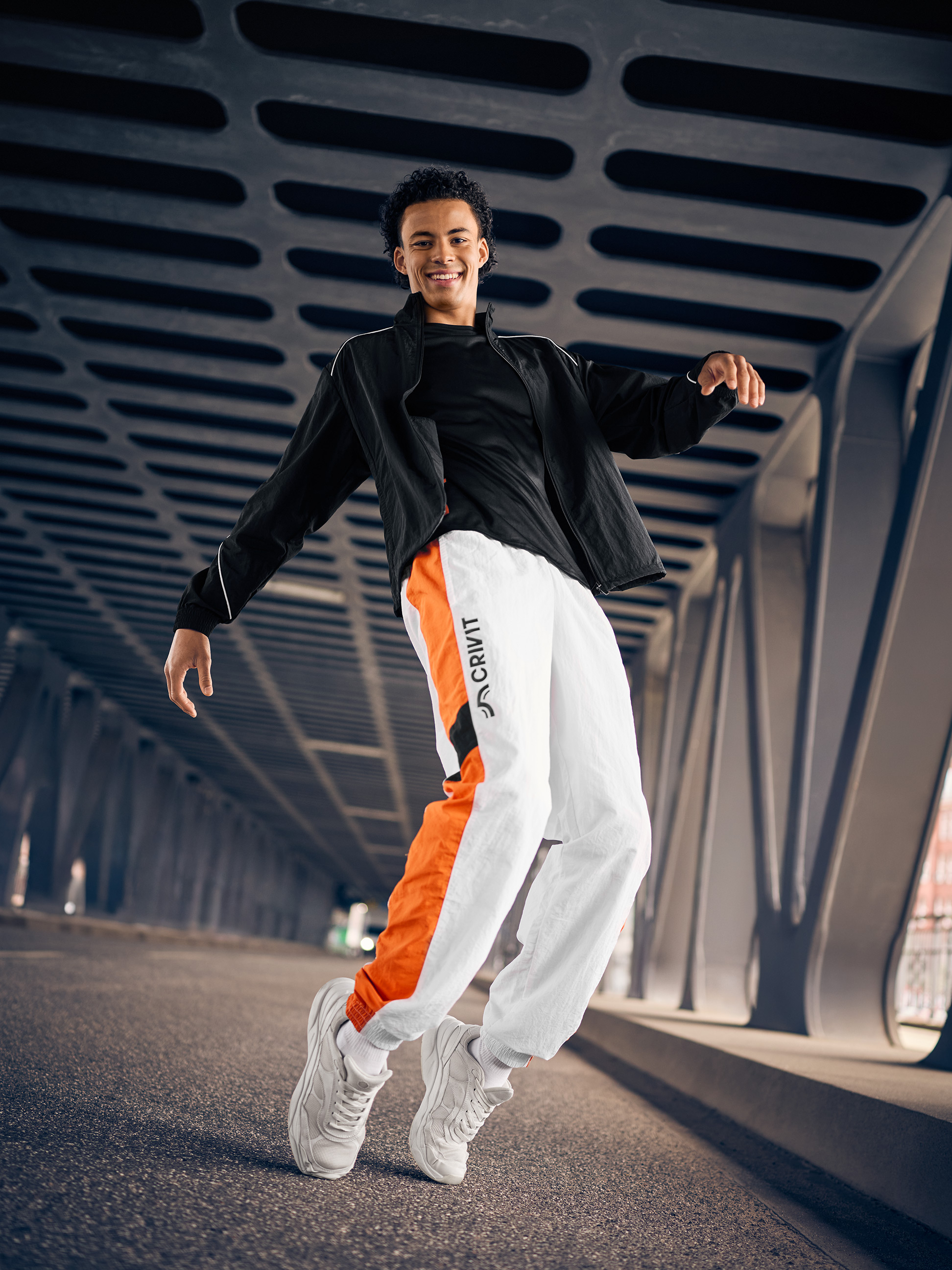 Mann in schwarzer Trainingsjacke und weißer Trainingshose mit orangefarbenen Details, springt auf einer Brücke.