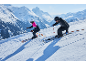 Zwei Skifahrer in Skibekleidung und Ausrüstung fahren den Berg hinunter.