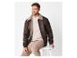 Mann in brauner Pilotenjacke mit cremefarbenem T-Shirt und beiger Cordhose, auf einem Hocker sitzend.