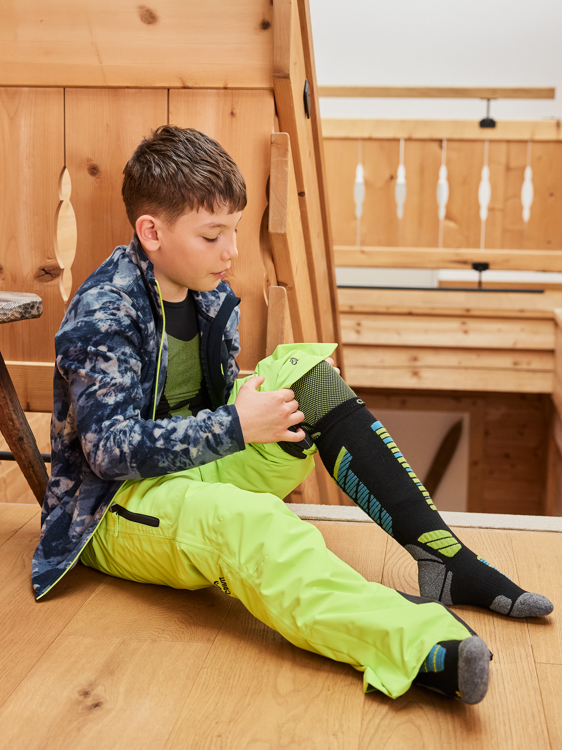 Sitzendes Kind in neongrüner Skihose und schwarzen Skisocken.