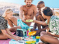 Eine Gruppe Menschen genießt ein Picknick am Strand mit einer Kühlbox und farbenfrohem Geschirr.