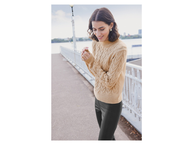 Eine Frau trägt einen beigen Strickpullover und schwarze Hosen.