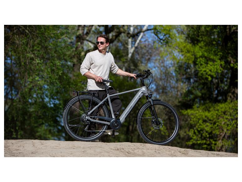 Mann in hellem T-Shirt und dunkler Hose, steht neben einem grauen E-Bike auf einem Feldweg.