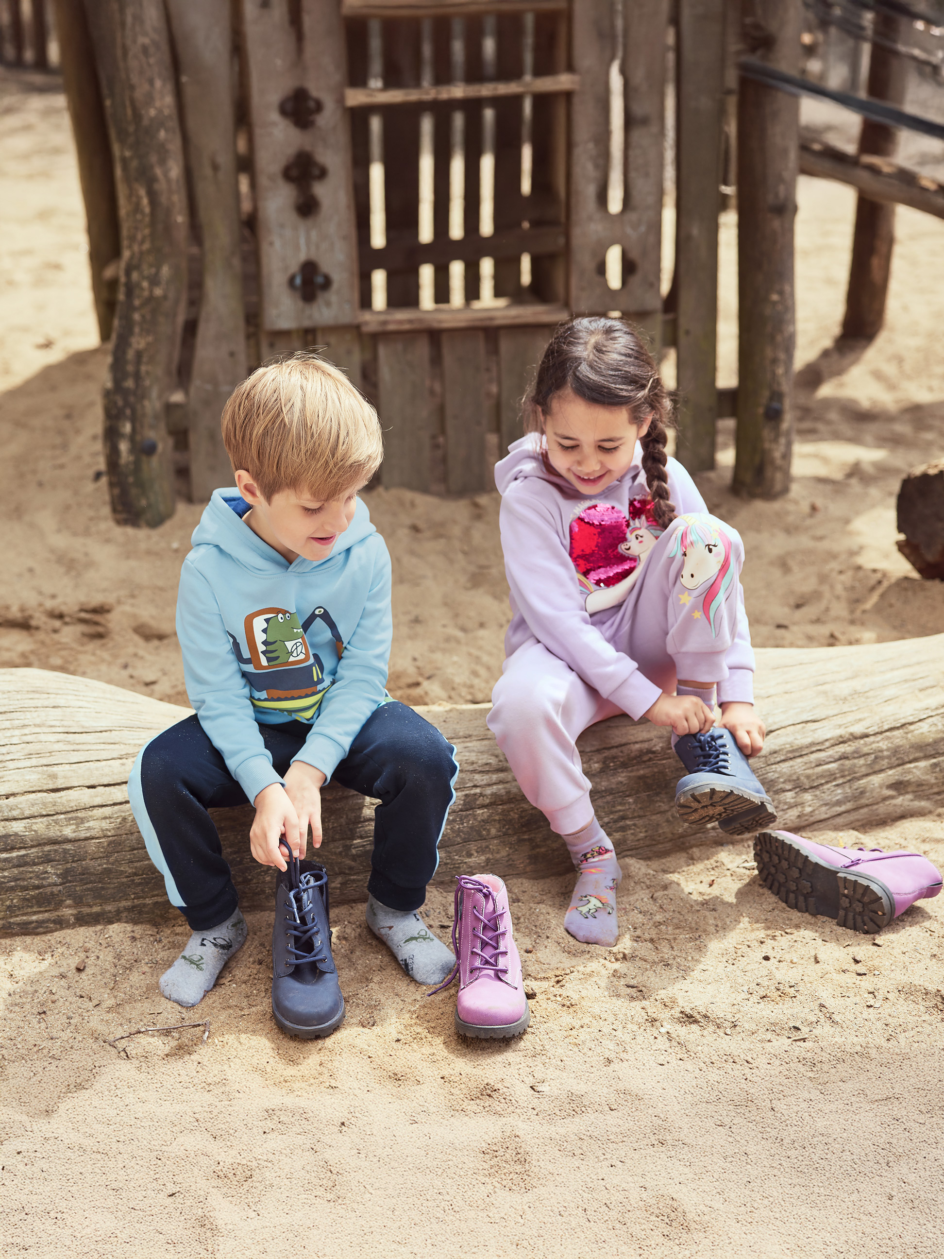 Zwei Kinder binden ihre Stiefel auf einem Baumstamm auf einem Sandspielplatz.