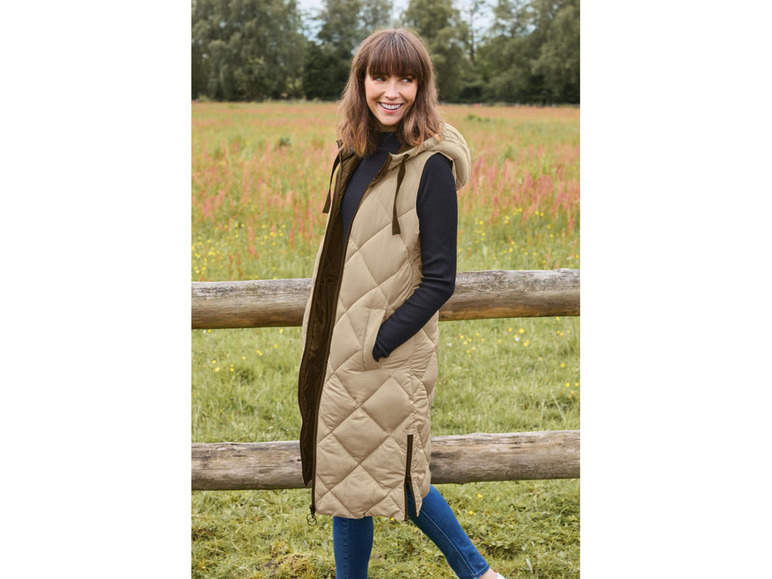 Eine Frau trägt eine lange, beige wattierte Weste mit Kapuze auf einem Feld.
