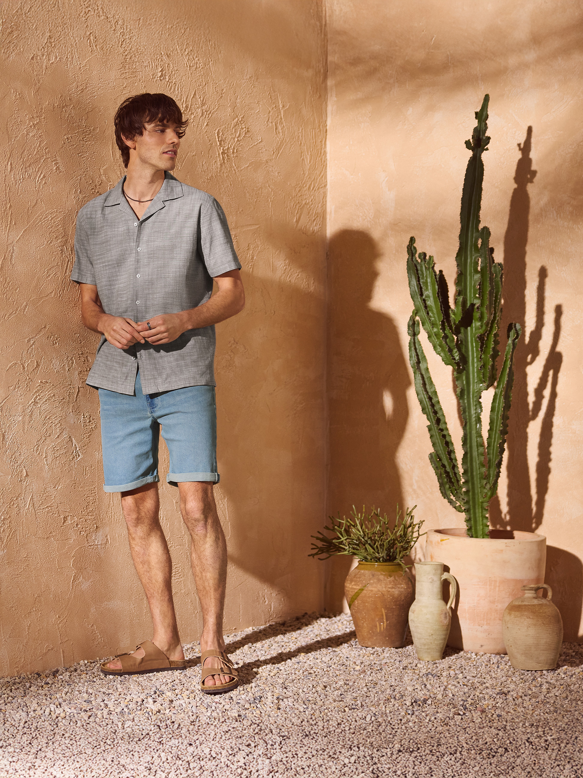 Mann in grauem Hemd und Jeans-Shorts, Sandalen, in mediterraner Umgebung.