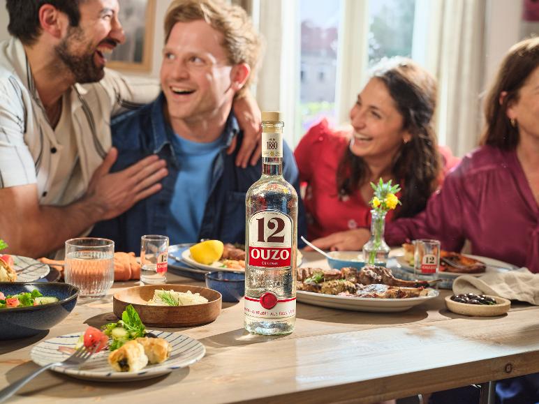 Eine Flasche Ouzo und Essen auf einem Tisch mit fröhlichen Menschen beim Abendessen.