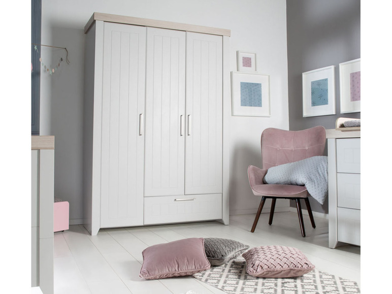 Ein weißer Kleiderschrank mit drei Türen und einer Schublade, ein rosa Sessel und Dekokissen.