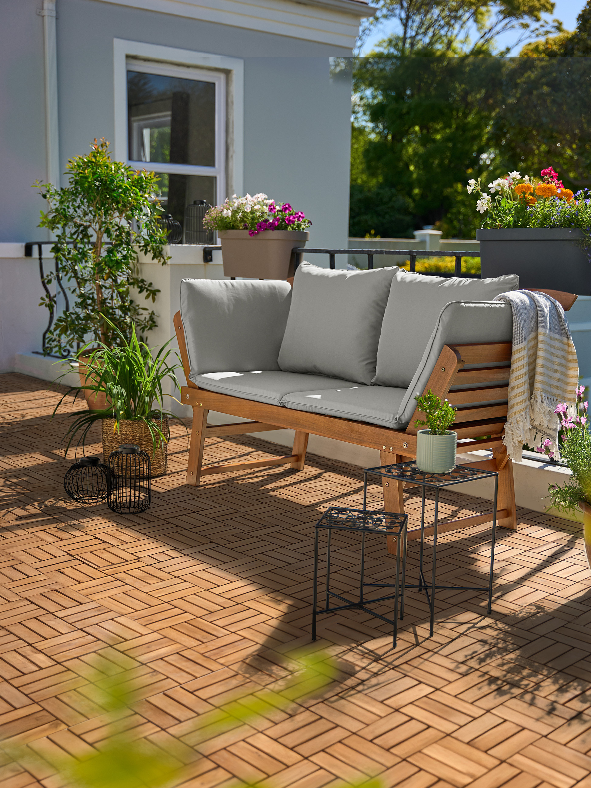 Gartensofa aus Holz mit grauen Kissen auf einem Balkon mit Holzfliesen und Pflanzen