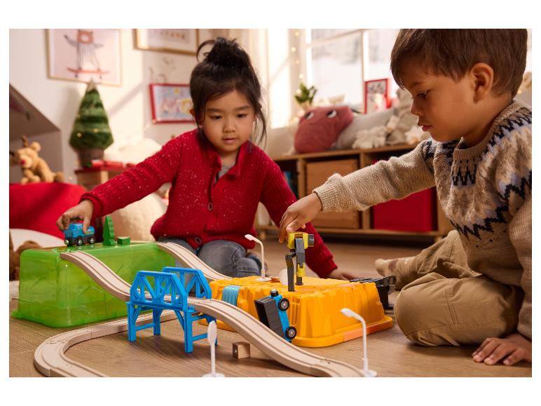 Zwei Kinder spielen mit einer Holzeisenbahn und Spielzeugautos auf dem Boden.
