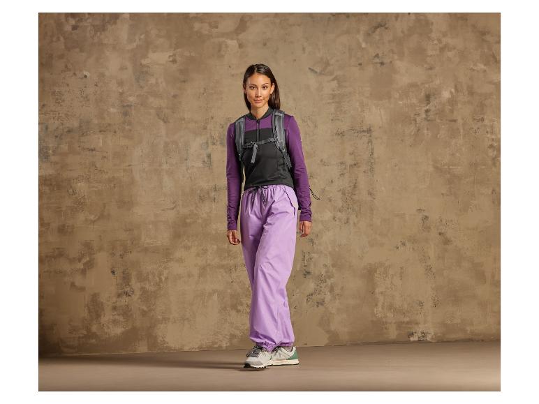 Eine Frau in lila Hose, schwarzer Weste und Sneakers vor einer rauen Wand.