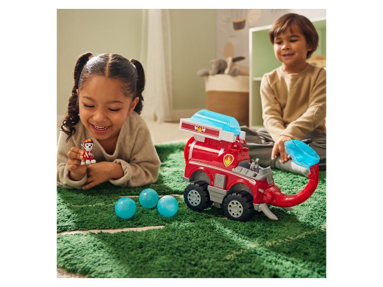 Zwei Kinder spielen mit einem Paw Patrol Fahrzeug und einer Figur auf einem grünen Teppich.