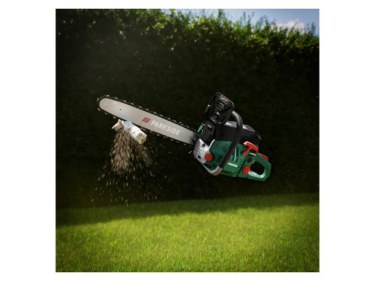Kettensäge schneidet einen Ast, mit Sägespänen in der Luft, vor einer grünen Hecke und Gras.