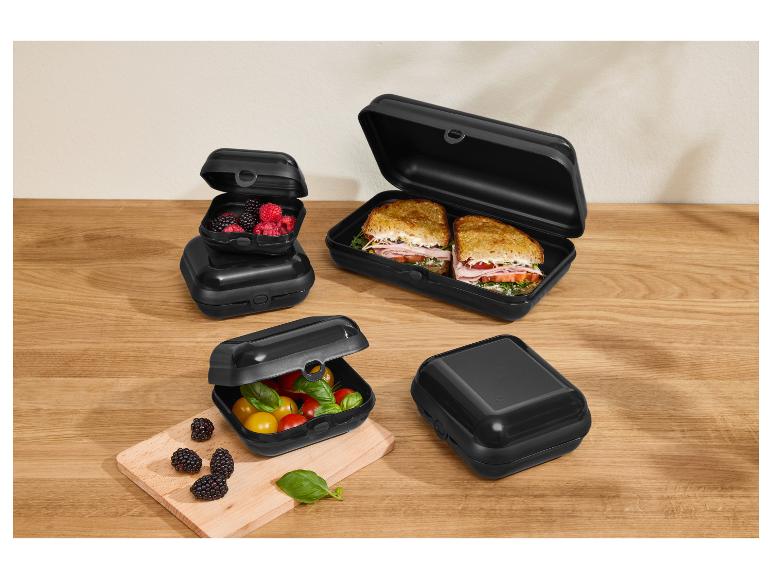 Schwarze Brotdosen mit Sandwiches, Beeren und Tomaten auf einem Holztisch.