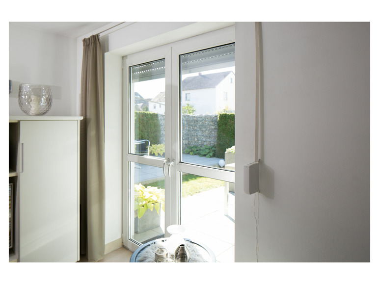 Ein weißer Motor für ein Rollladen, montiert an einer weißen Wand, neben einem französischen Balkon mit Blick auf einen grünen Garten.