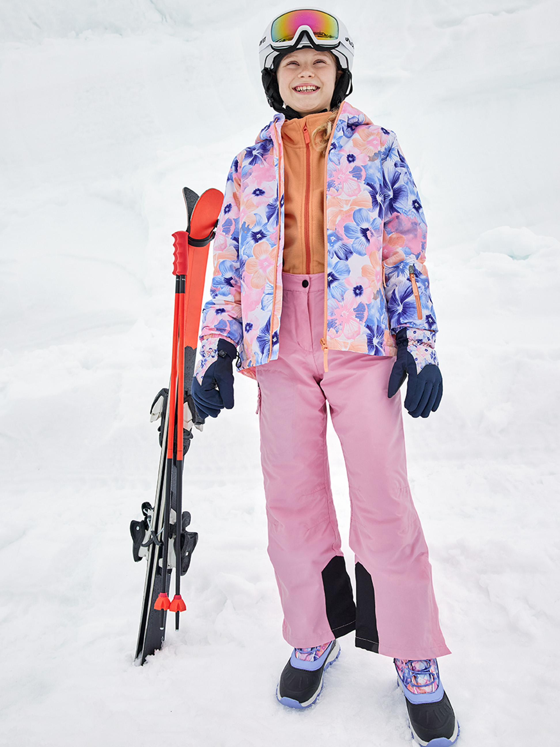 Lächelndes Mädchen in floraler Skijacke, rosa Hose und Helm, mit Skiern.