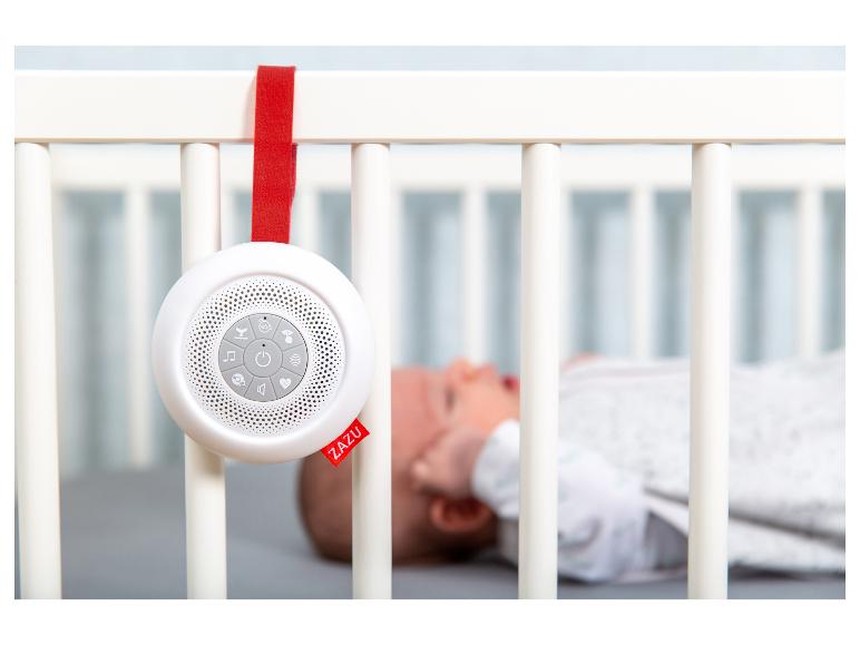 Weiße ZAZU Soundmaschine hängt an einem Babybett mit schlafendem Baby im Hintergrund.