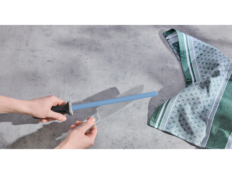 Hände schärfen ein Messer mit einem blauen Wetzstahl, daneben ein grün-weißes Küchentuch.