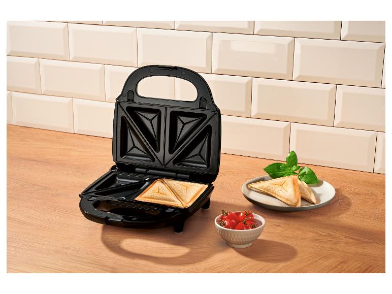 Sandwichmaker mit fertigen Sandwiches, Tomaten und Basilikum auf einer Holzoberfläche.