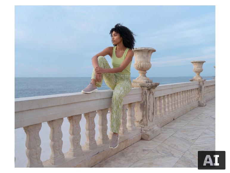 Eine Frau in einem hellgrünen ESMARA® Top und einer gemusterten Hose sitzt auf einem Geländer am Meer. [KI-generiertes Bild]