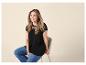 Lächelnde Frau in schwarzem Spitzen-T-Shirt und Jeans, auf einem Hocker sitzend.