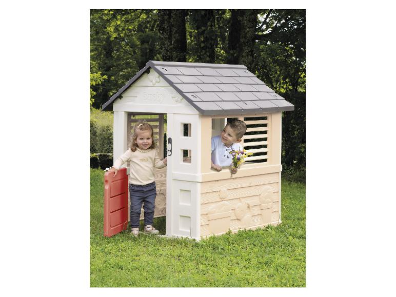 Zwei Kinder spielen in einem Plastik-Spielhaus mit grauem Dach und roter Tür in einem Garten.