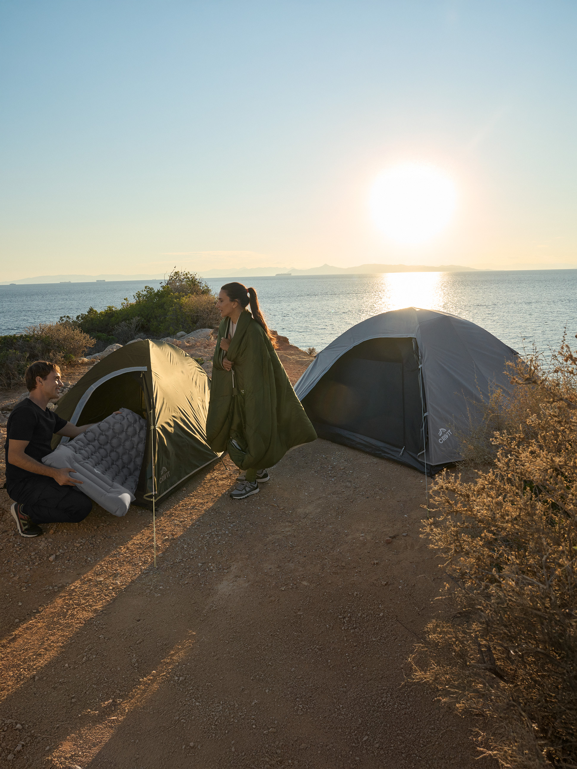 Zwei Personen campen am Meer mit zwei Crivit Zelten und einer Luftmatratze.