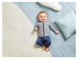 Ein Baby in einer grauen Jacke und blauen Hosen liegt auf einer weißen Decke mit blauen Kissen.