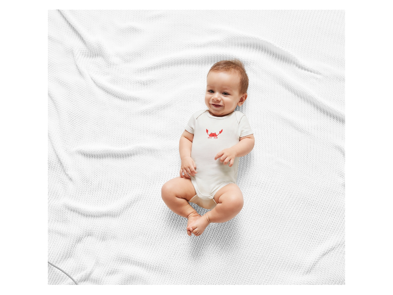 Lächelndes Baby in weißem Body mit Krabbenmotiv auf einer weißen Decke.