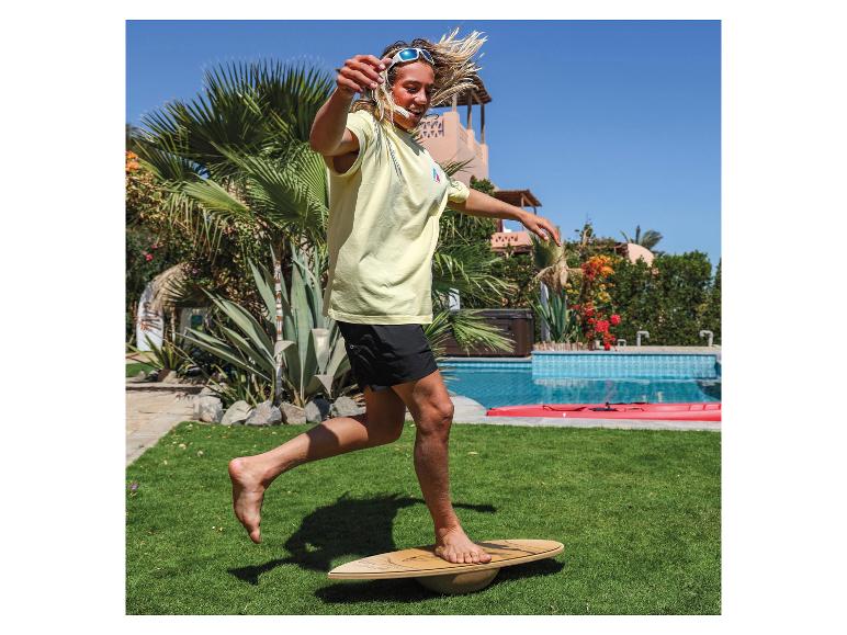 Mann balanciert auf einem Holz-Balanceboard auf einem sonnigen Rasen neben einem Pool.