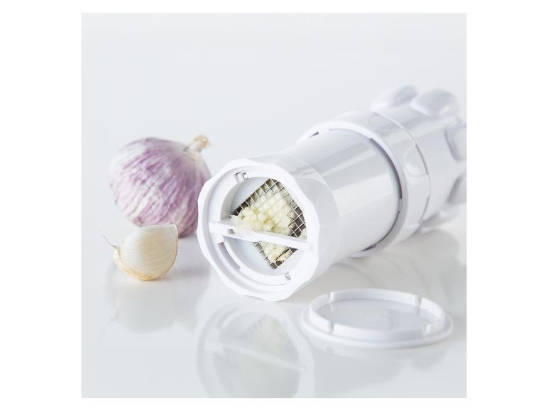 Weiße Knoblauchpresse mit gehacktem Knoblauch und einer Knoblauchzehe auf weißer Oberfläche.