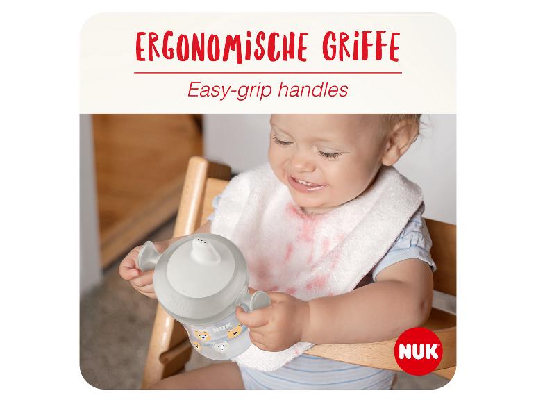 Lächelndes Baby mit verschmiertem Lätzchen, das einen Trinklernbecher mit ergonomischen Griffen hält.