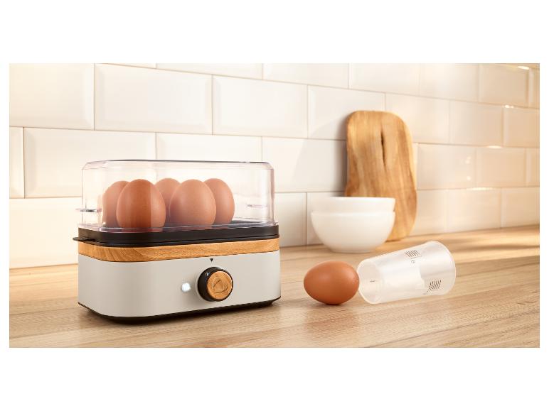 Eierkocher mit Holzoptik, Eier und Messbecher auf einer Küchenarbeitsplatte.