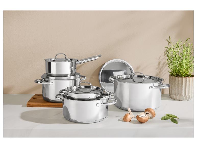 Set aus glänzenden Edelstahl-Töpfen und -Pfannen auf einer Küchenarbeitsplatte mit Pilzen und Kräutern.