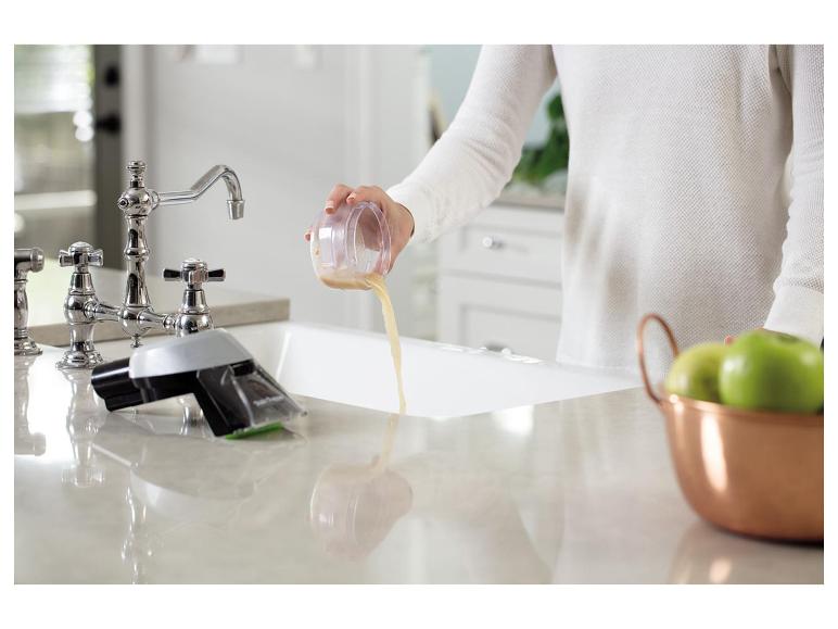 Eine Person gießt Flüssigkeit aus einem Behälter in ein Spülbecken, mit grünen Äpfeln daneben.