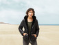 Eine Frau trägt eine schwarze Jacke mit Kapuze am Strand.