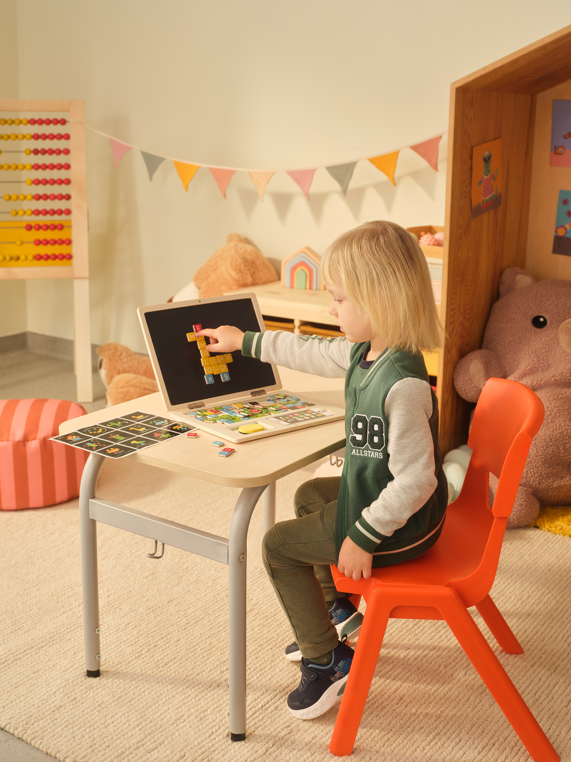 Kind spielt mit einem Holzlaptop und Magnetbuchstaben an einem kleinen Schreibtisch.