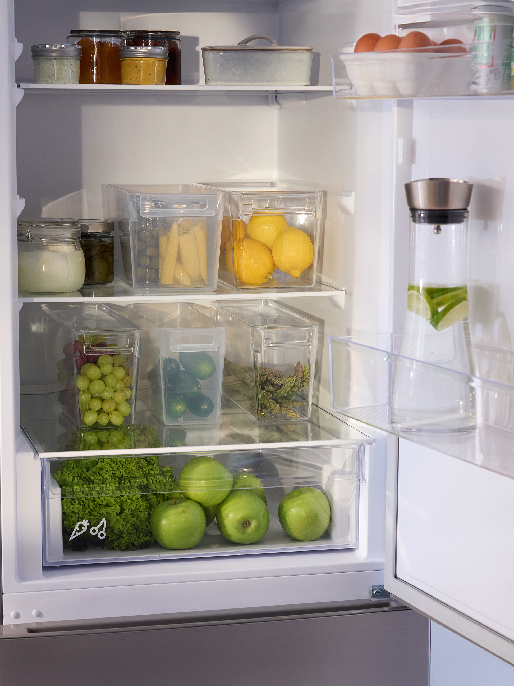 Offener Kühlschrank mit organisierten durchsichtigen Behältern für frische Produkte, Saucengläsern und einem Wasserkrug.