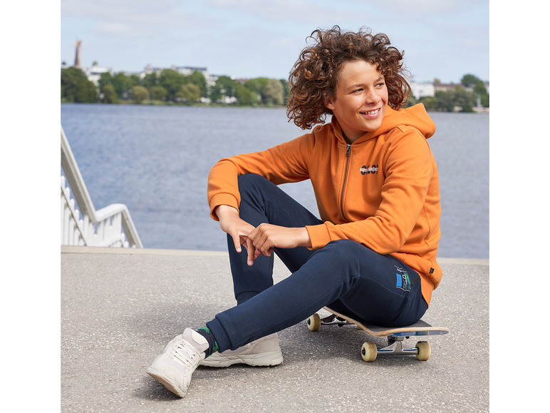 Ein Junge in einem orangenen Hoodie und blauen Jogginghosen sitzt auf einem Skateboard.