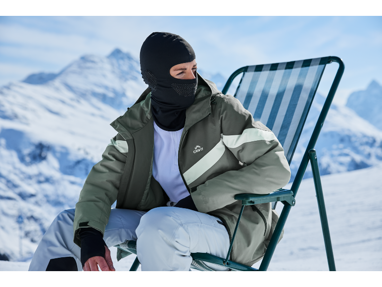 Mann mit Sturmhaube und grüner Skijacke sitzt auf einem Stuhl in verschneiten Bergen.