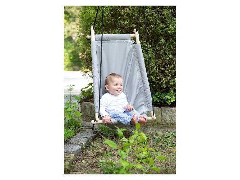 Glückliches Kleinkind sitzt in einer grauen Stoffschaukel, die draußen hängt.
